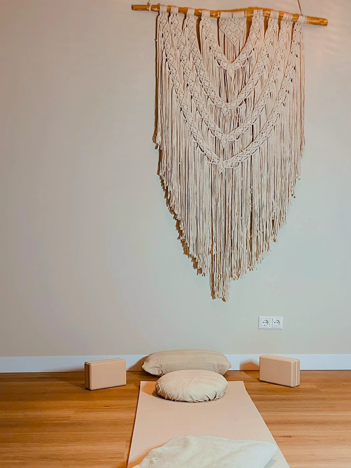 Een mat met kussen en kurkblokken op de vloer, onder een macramé wandkleed